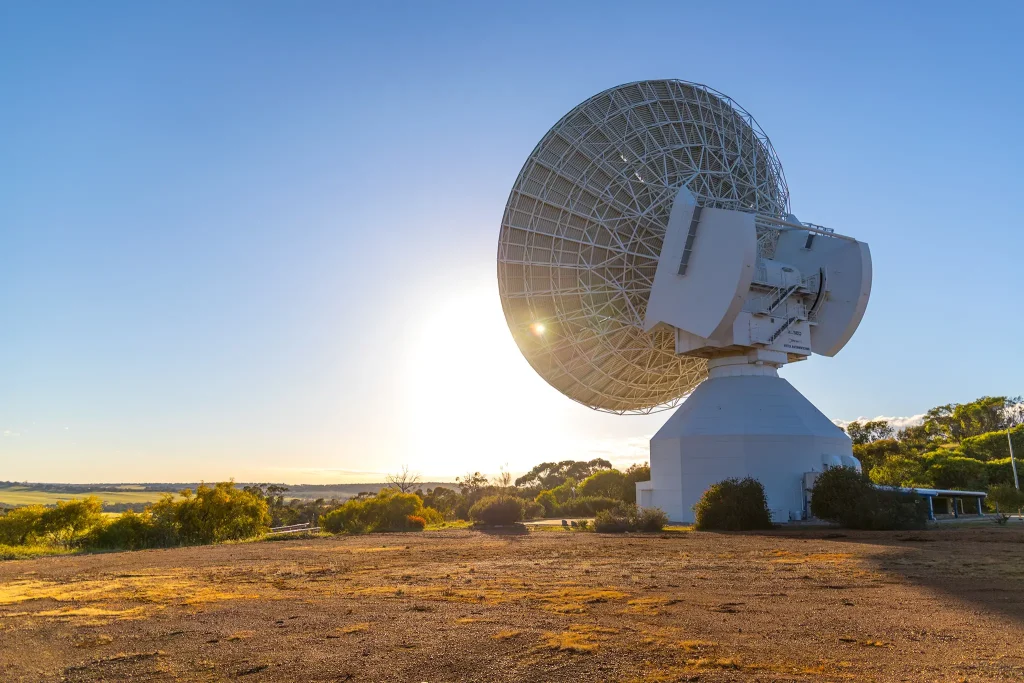 Large radio telescope antenna facing sky at sunset, with sun flare and clear blue sky in background, situated in a dry landscape.