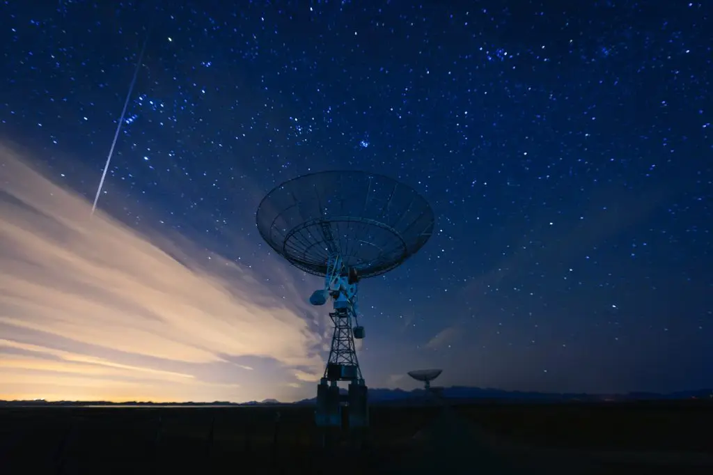 A satellite dish under a starry sky with a shooting star and wispy clouds, during twilight.