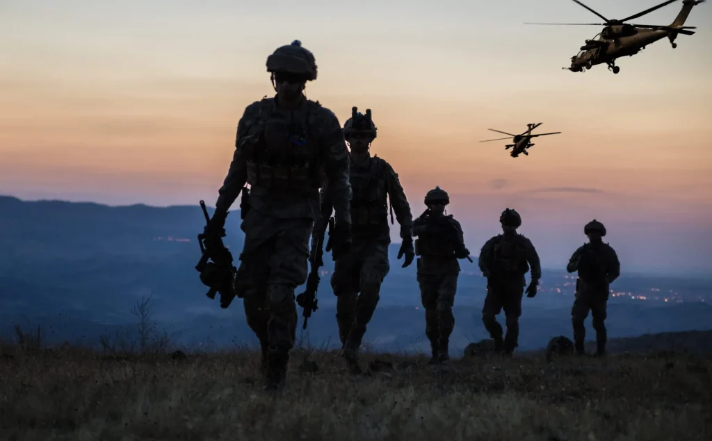 Soldiers walking in a line at dusk with a helicopter flying in the background.
