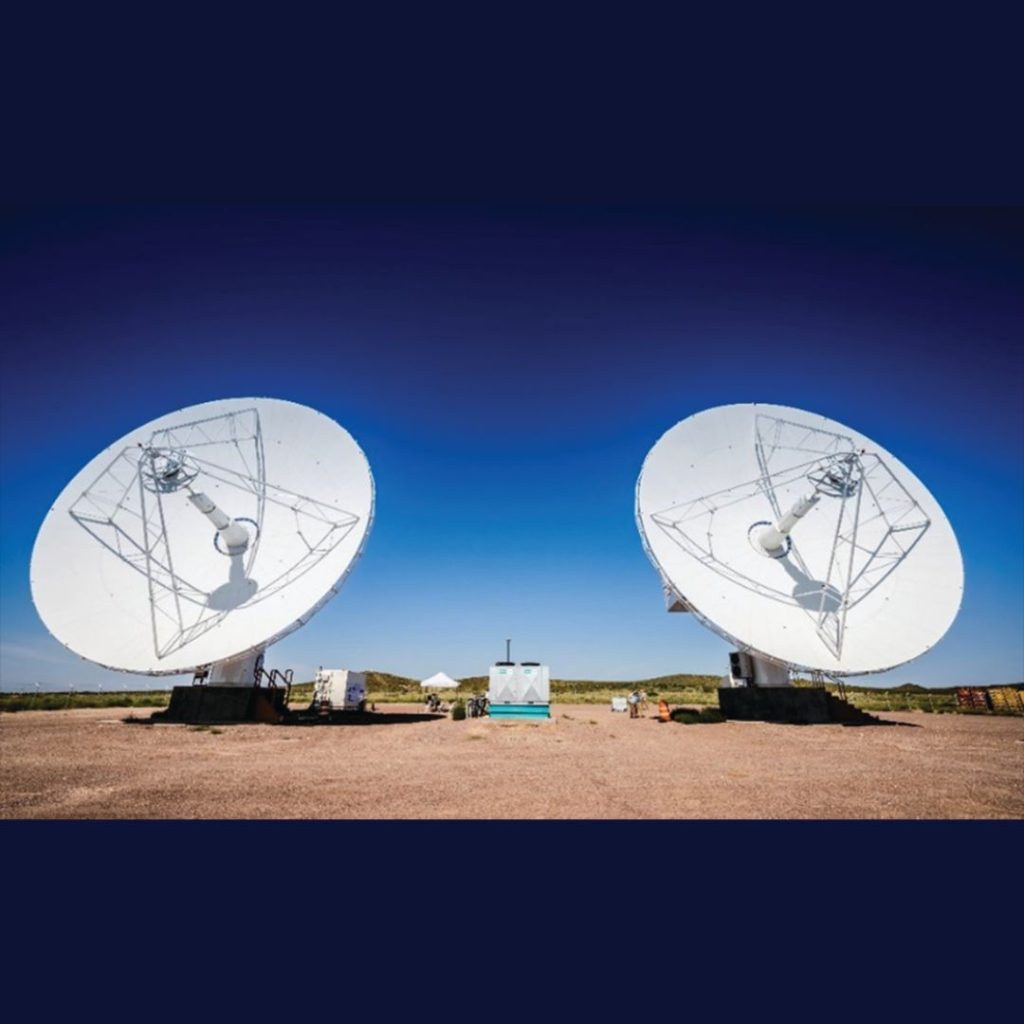 Two large satellite dishes are aligned and facing skyward in an open, barren ground setting under a clear, blue sky.