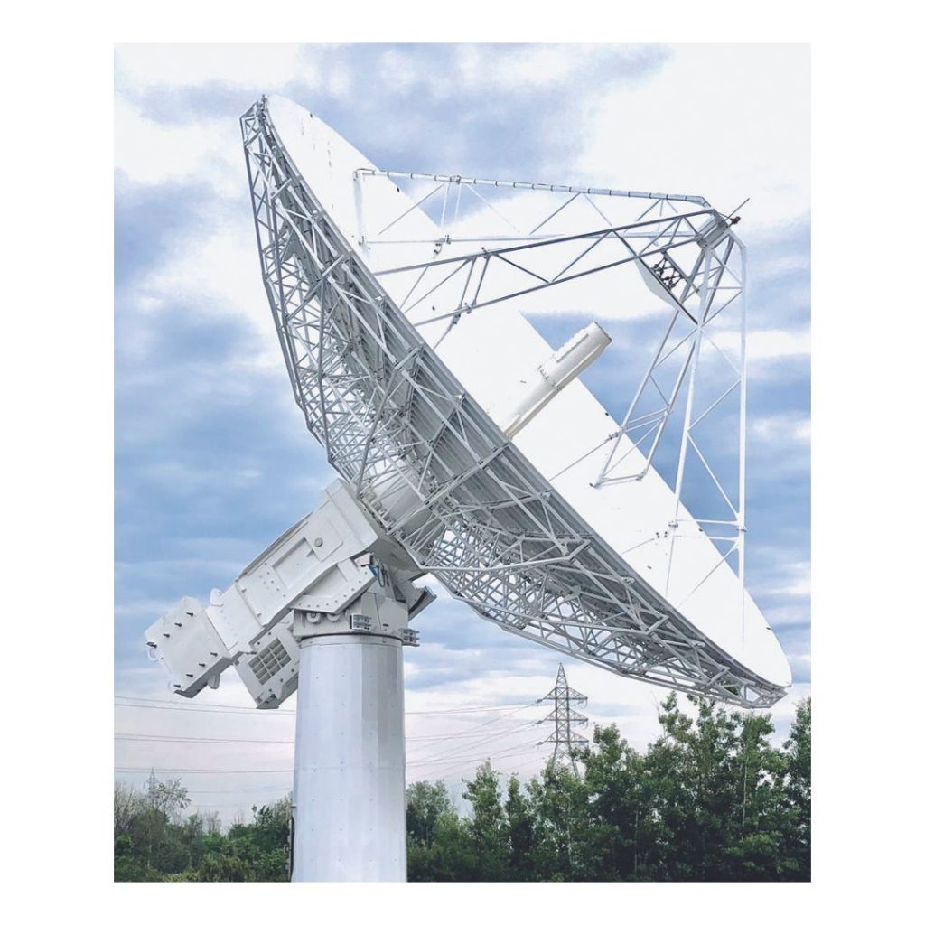 A large, white satellite dish antenna is mounted on a tall steel structure against a partly cloudy sky, with trees and a transmission tower in the background.