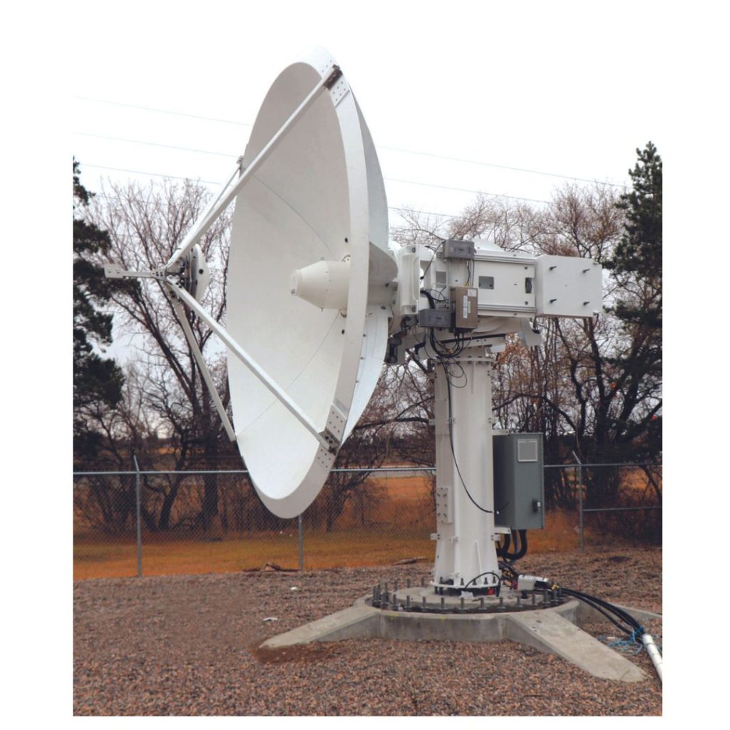 A large white satellite dish is mounted on a sturdy base in an outdoor area. The background includes bare trees and a chain-link fence.