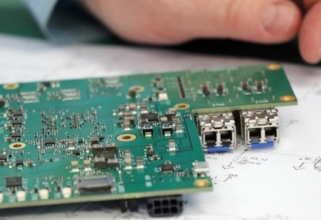 A close-up of a person’s hand next to a green printed circuit board with various electronic components, including USB ports, placed on technical drawings.