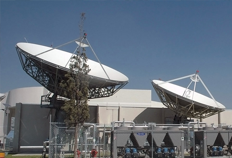 Two large satellite dishes within a secure, fenced area, with industrial buildings and clear sky in the background.