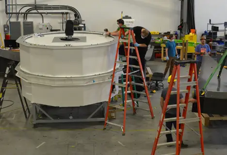 Plusieurs personnes travaillent autour d'un grand équipement cylindrique à l'aide d'échelles dans un atelier industriel.