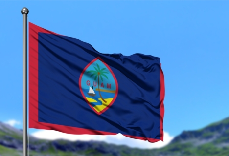 The flag of Guam with a blue field, red border, and the central emblem featuring a coconut tree, a sailboat, and the word "GUAM," flutters in the wind against a blue sky and mountainous background.