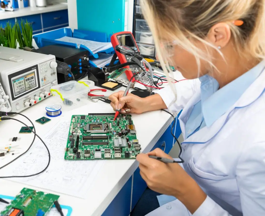 A person in a lab coat is soldering a component on a green circuit board in an electronics lab, surrounded by various electronic tools and equipment.