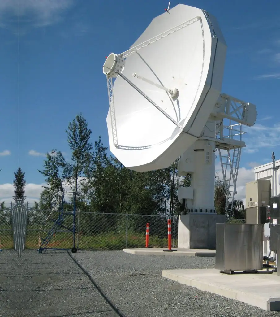 Large satellite dish at a ground station on a sunny day, surrounded by a chain-link fence and trees under a clear sky.
