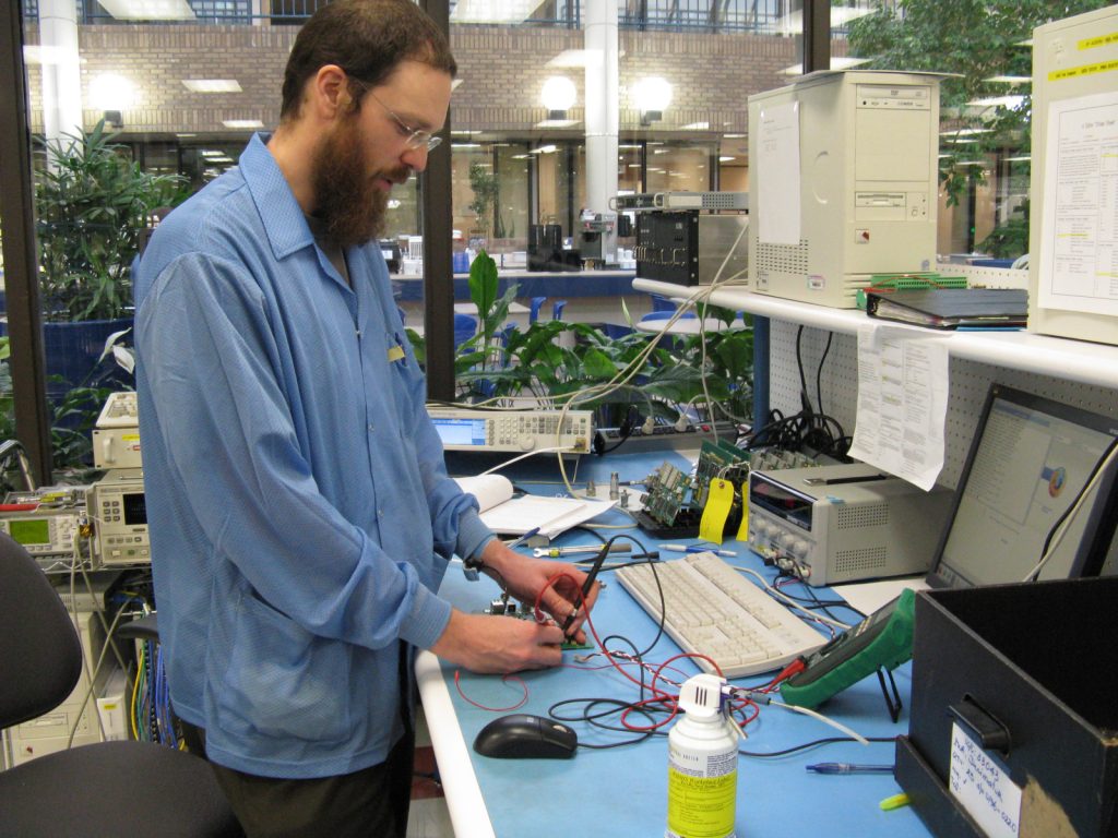 A person with a beard is standing at a workbench in an electronics lab, using multimeter probes on a circuit board. The desk is cluttered with electronic equipment, a computer, and a can of cleaner.