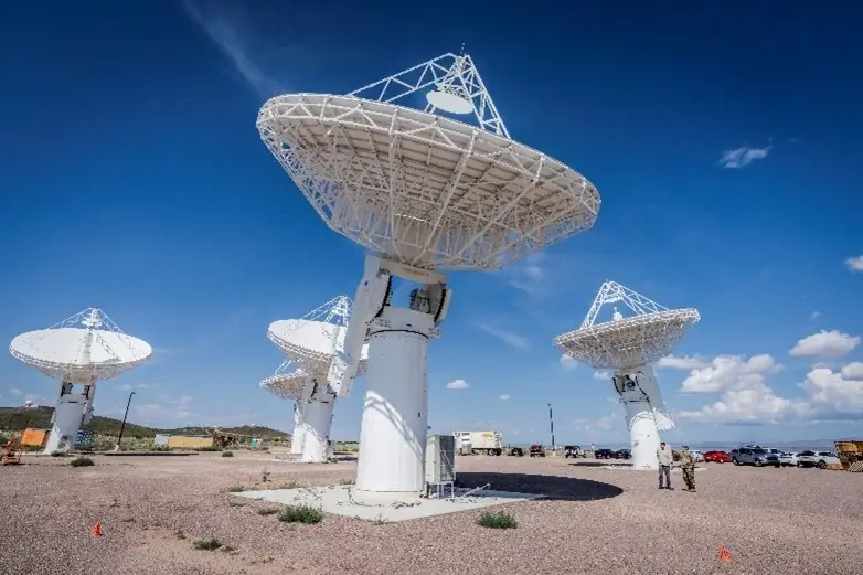 A row of large white satellite dishes facing the sky, set against a backdrop of blue sky with clouds, in a remote desert-like area.