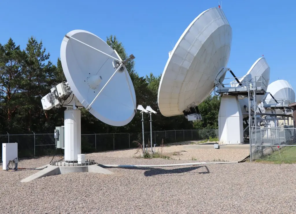 Several large satellite dishes are positioned outdoors in a fenced area, pointed towards the sky, with trees and clear blue sky in the background.