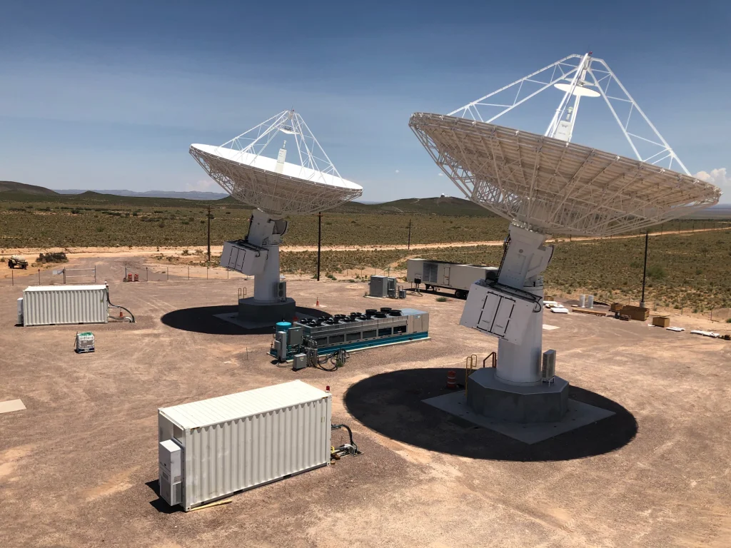 Three large satellite dishes at a remote desert facility under a clear blue sky.