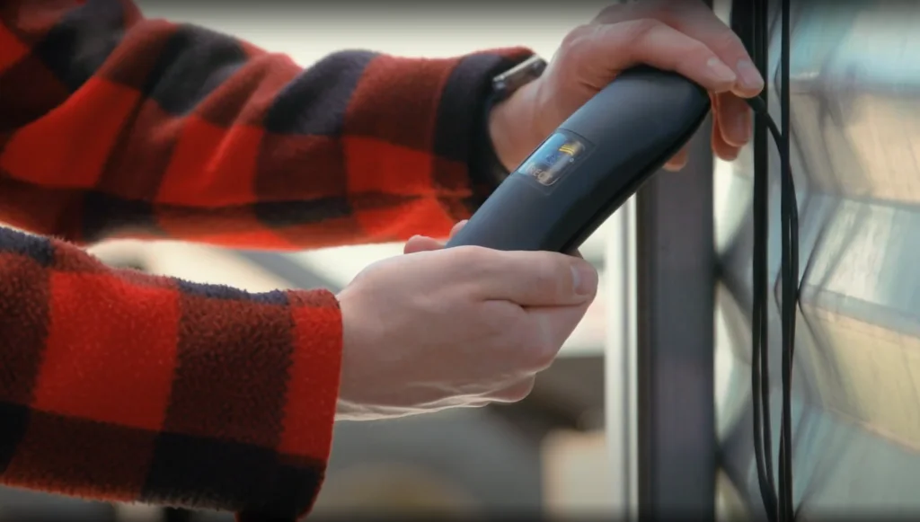 A person in a red and black checkered shirt holds a handheld scanning device next to a window, examining corded wires hanging from it.