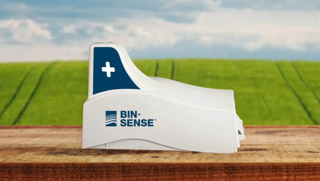 A white device labeled "BIN-SENSE" with a blue cross symbol, placed on a wooden surface, with a background of a grassy field and cloudy sky.