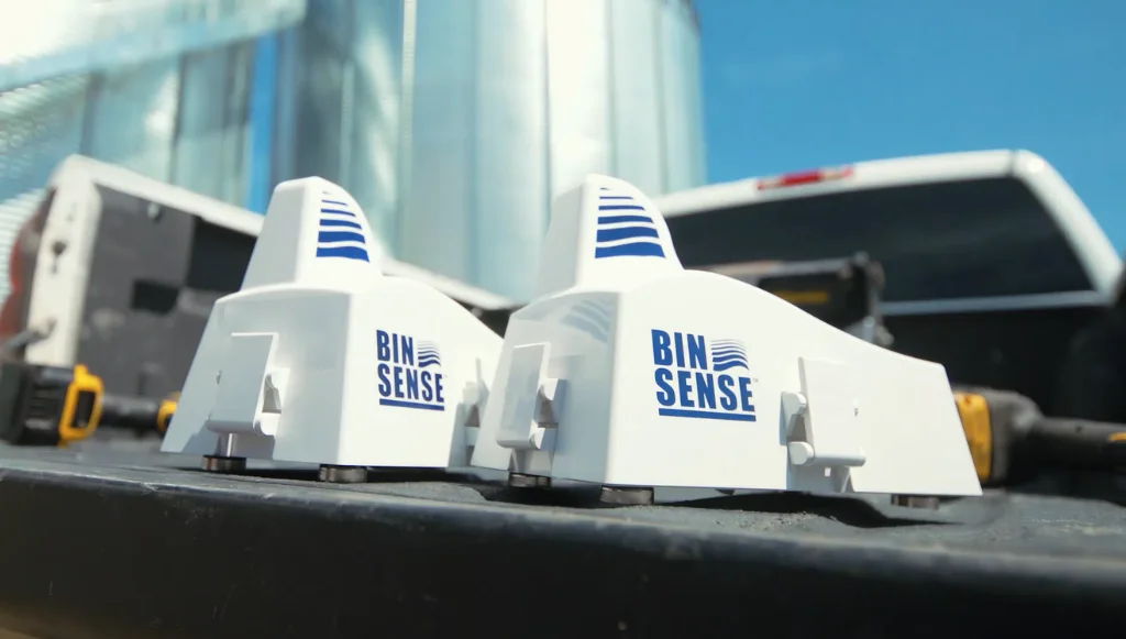 Two Bin Sense devices are placed on the back of a truck bed. The devices are white with blue text and logos, and there is farming equipment and a silo in the background.