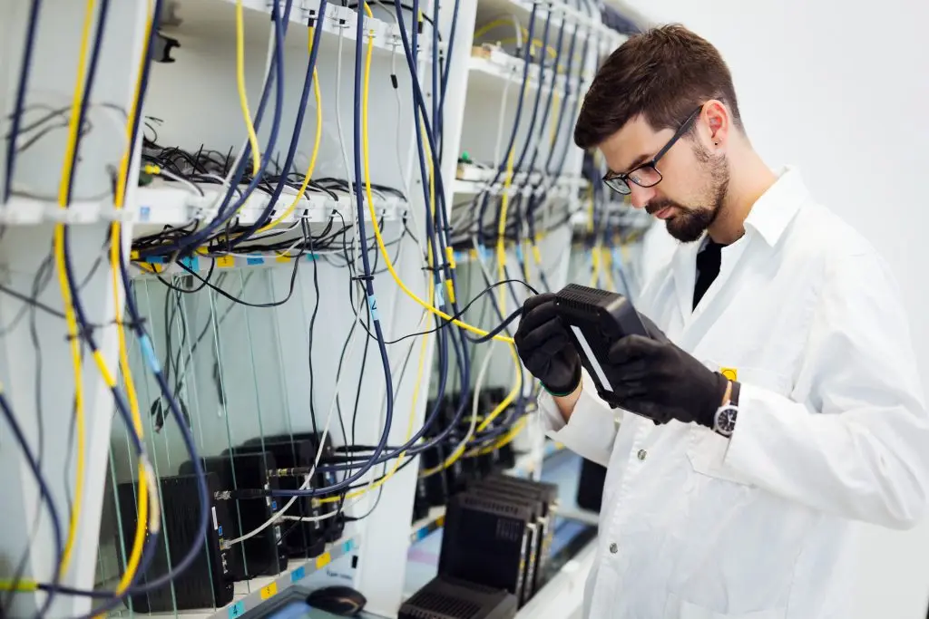 A male technician in a lab coat and glasses examines network equipment with a handheld device in a server room.