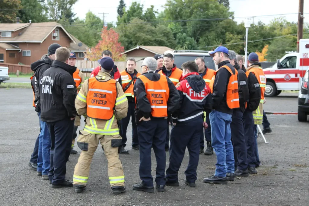 A group of emergency responders in uniforms, including firefighters and decontamination officers, gather for a briefing in a parking lot.