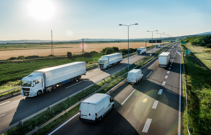 Several trucks and vans are traveling on a multi-lane highway surrounded by open fields under a clear sky.