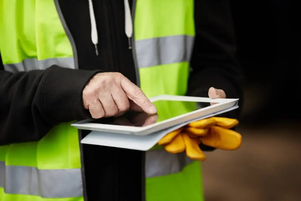 A person in a high-visibility vest uses a tablet while holding a pair of gloves.