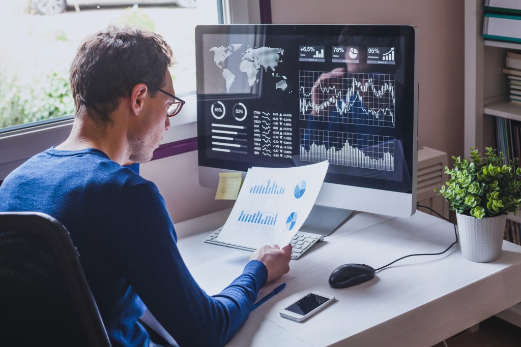 Un homme vêtu d'un pull bleu examine des documents financiers, avec devant lui un écran d'ordinateur affichant des graphiques et des données analytiques.