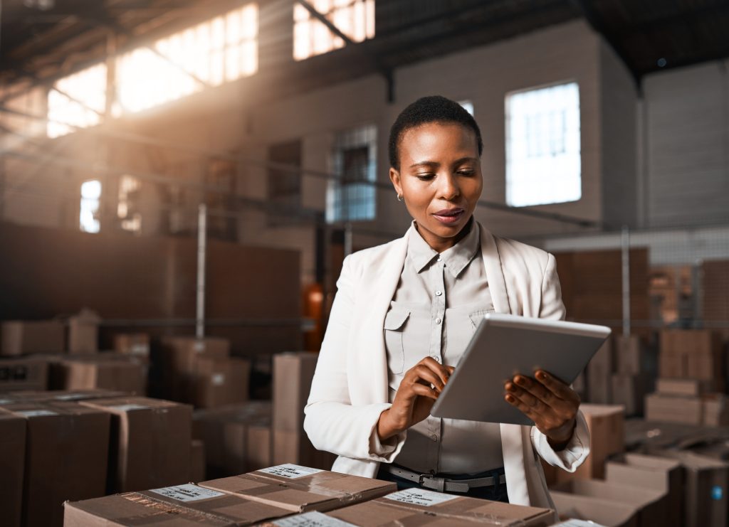 A person in a light-colored suit jacket uses a tablet while standing among boxes in a warehouse.