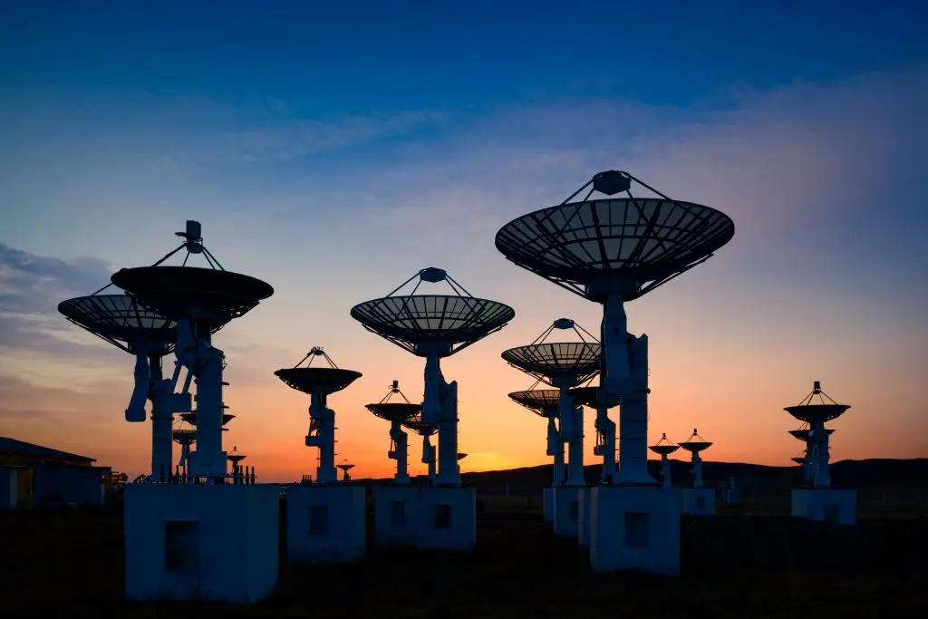 Silhouettes of multiple large satellite dishes at sunset. The dishes are uniformly spaced and aimed upwards against a colorful evening sky.