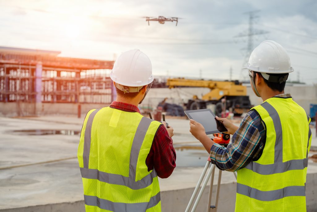 Two construction workers wearing hard hats and reflective vests operate a drone and use a tablet on a construction site with a partially built structure and heavy machinery in the background.