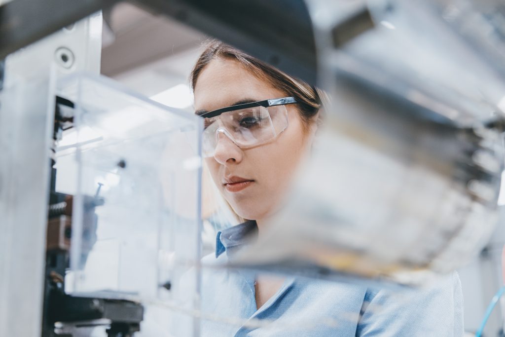 A woman wearing safety glasses attentively examines machinery in a high-tech laboratory.