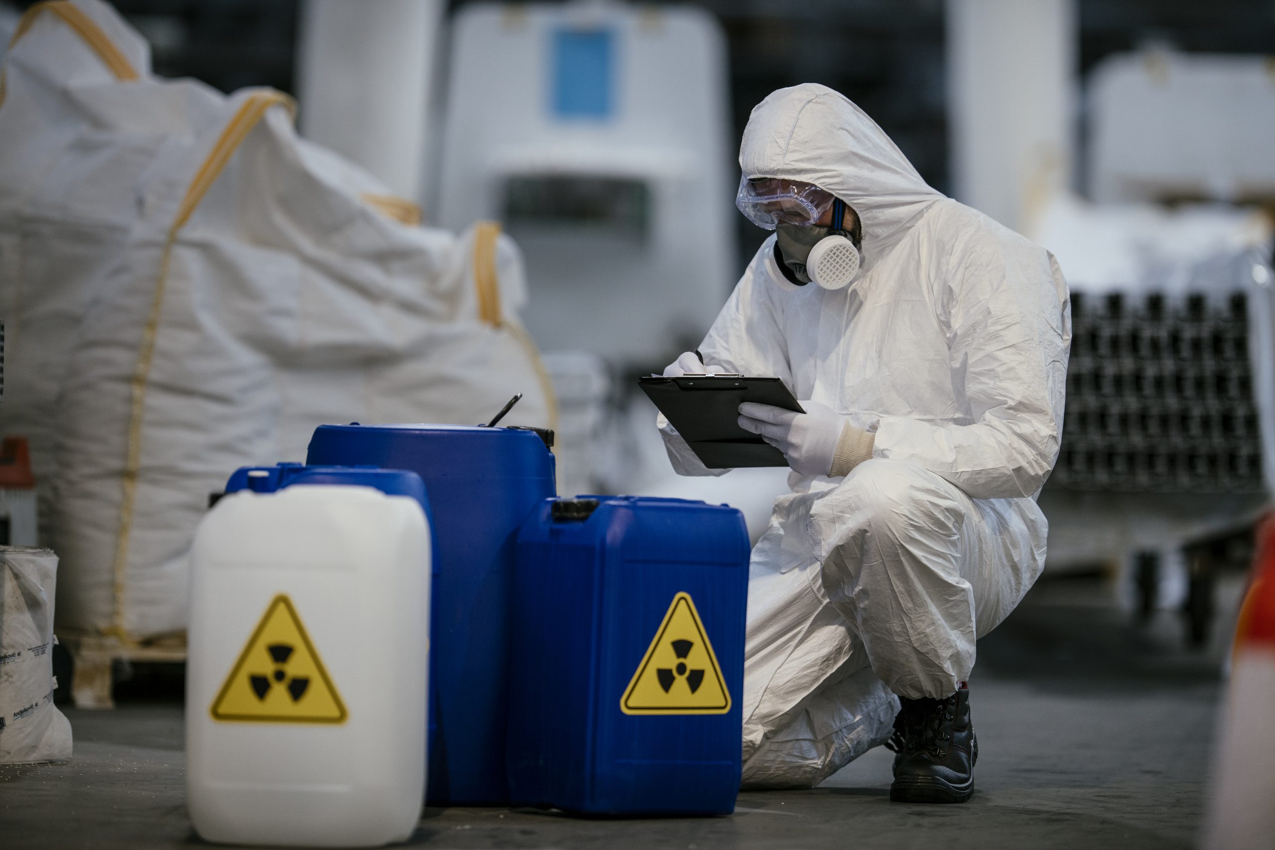 A worker in a protective suit and mask inspects a digital tablet while sitting beside blue chemical containers marked with hazard symbols in a warehouse.