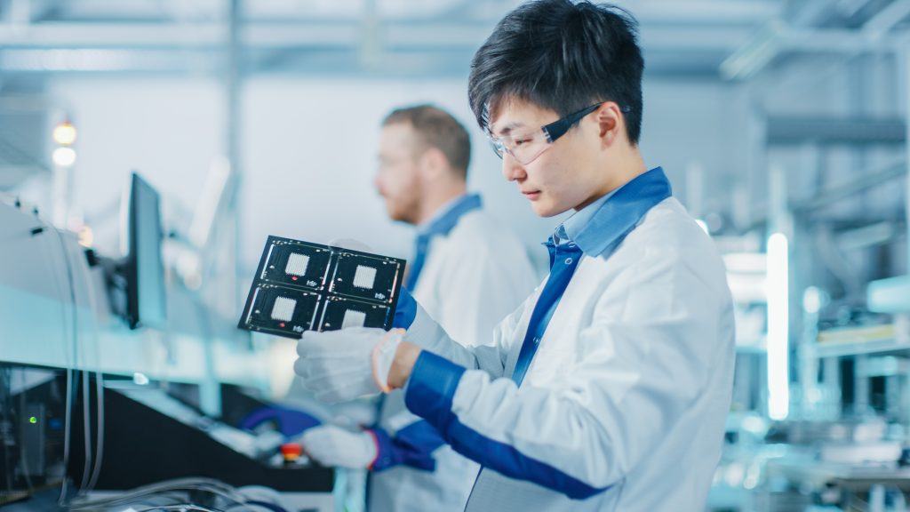 Two engineers, one Asian and one Caucasian, examine electronic components in a high-tech laboratory.