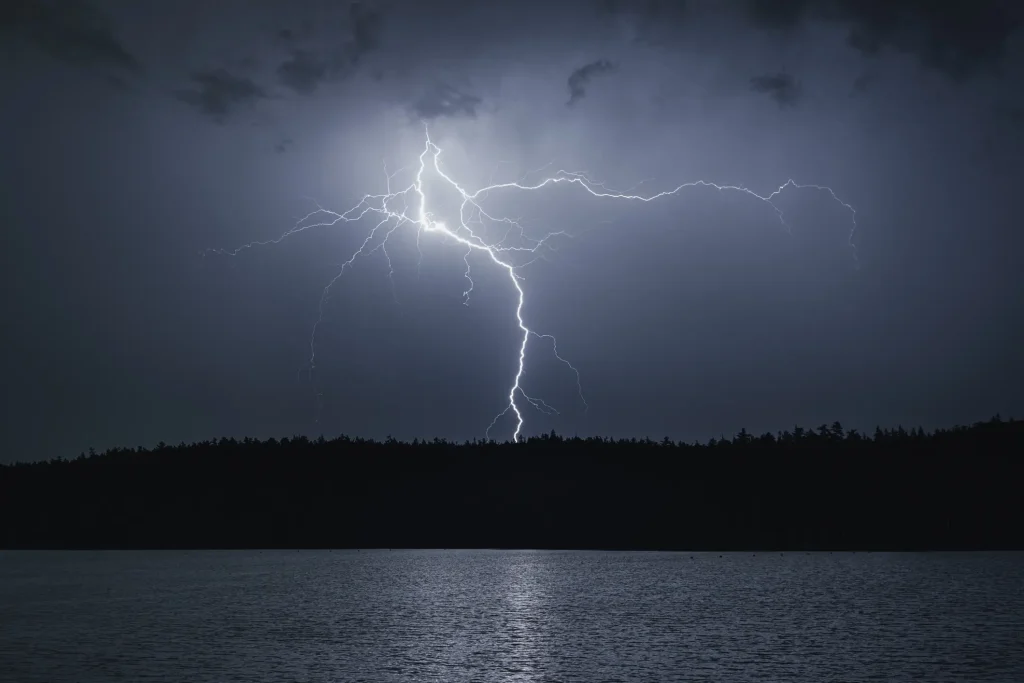 Lightning striking over a lake at night with dark silhouetted trees along the shoreline and a cloudy sky.