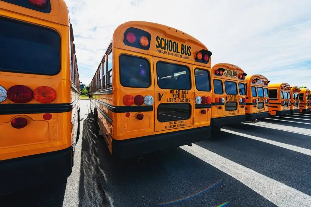 A row of parked yellow school buses seen from the rear, with safety signs and tail lights visible, under a partly cloudy sky.