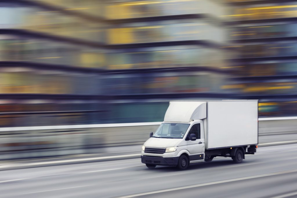 A white delivery truck drives quickly on a city street with blurred office buildings in the background.