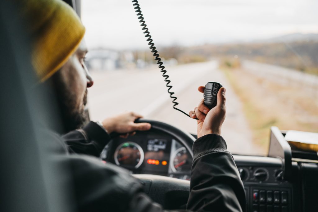 A person wearing a yellow beanie is driving a vehicle and holding a CB radio microphone, with a view of the road ahead through the windshield.