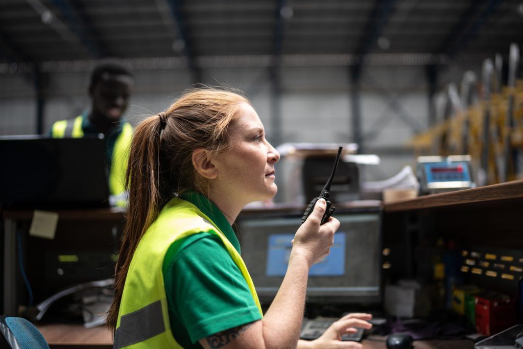 Une femme portant un gilet de sécurité jaune parle dans un talkie-walkie à un poste de travail dans un entrepôt. Une autre personne portant un gilet de sécurité est visible à l'arrière-plan.