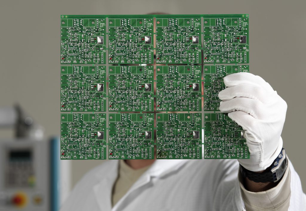 Un technicien en blouse de laboratoire, portant des gants blancs, tient un grand panneau de cartes de circuits imprimés vertes.