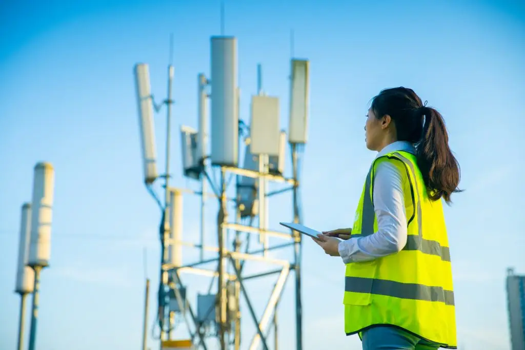 A woman in a high-visibility vest holds a tablet while looking at cellular antennas on a tower.