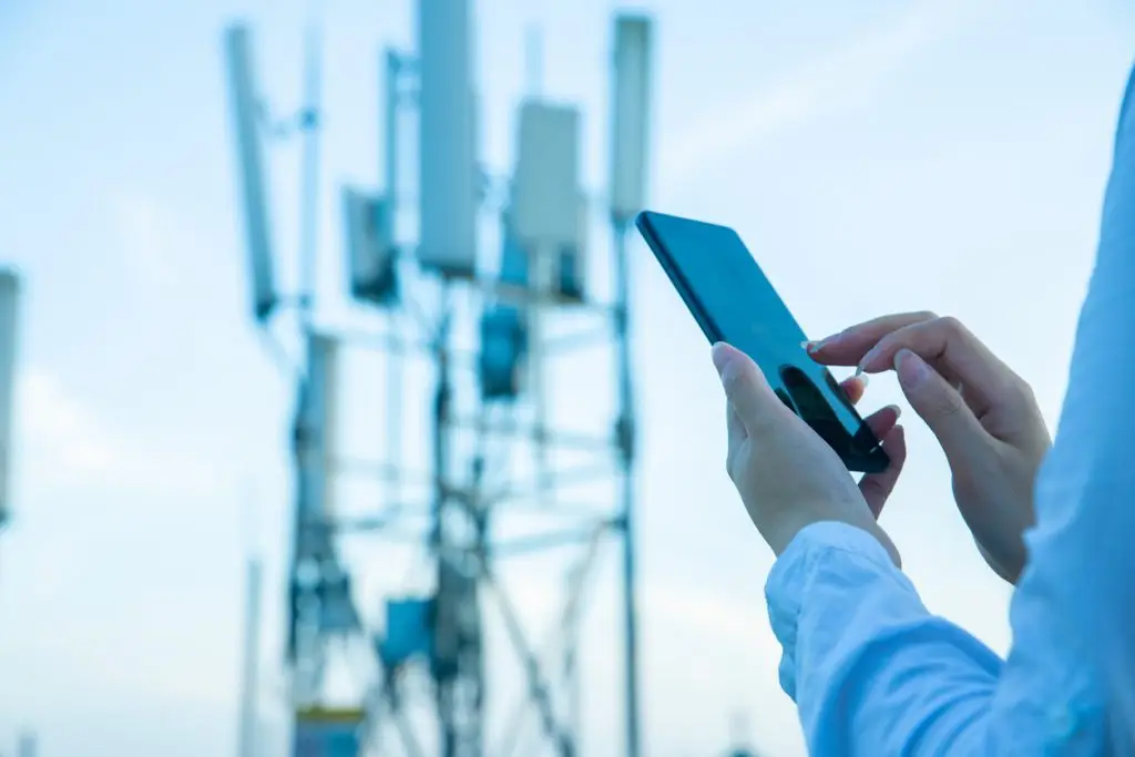 A person holding and using a smartphone with a blurred telecommunications tower in the background.