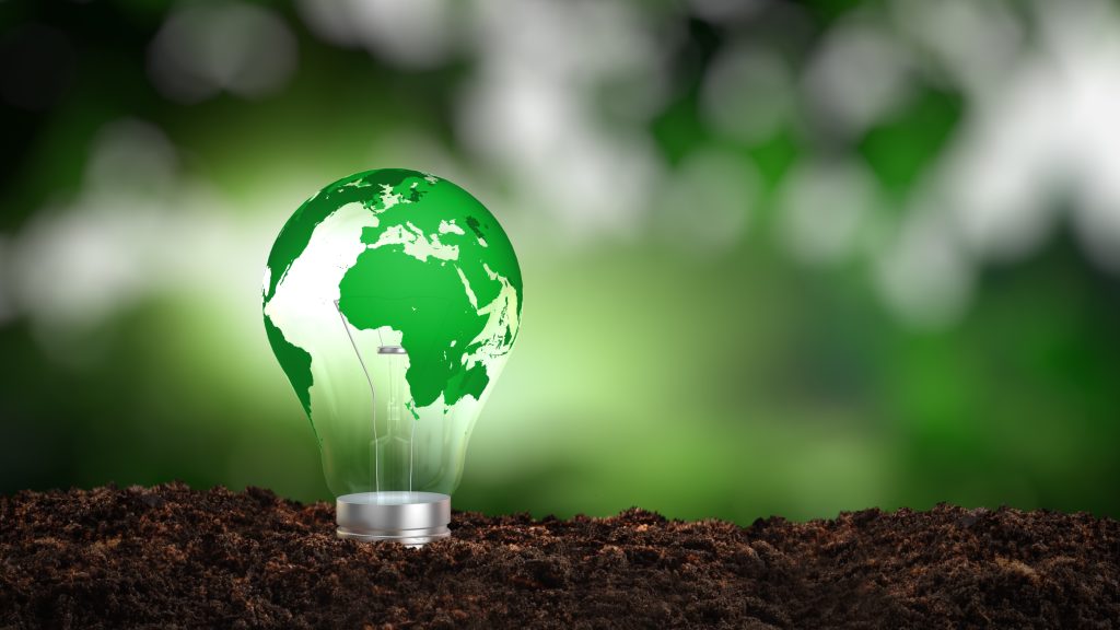 A green, earth-shaped lightbulb stands on soil against a blurred green backdrop, symbolizing eco-friendly energy solutions.
