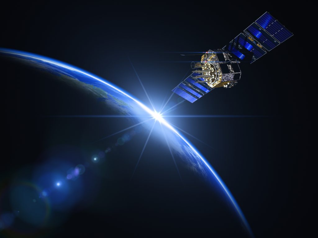 A satellite with extended solar panels orbits earth, illuminated by the sun against the dark backdrop of space.