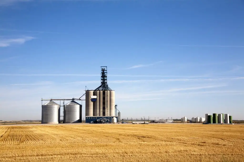 A large agricultural complex with multiple silos sits in the middle of an expansive, harvested field under a clear blue sky.