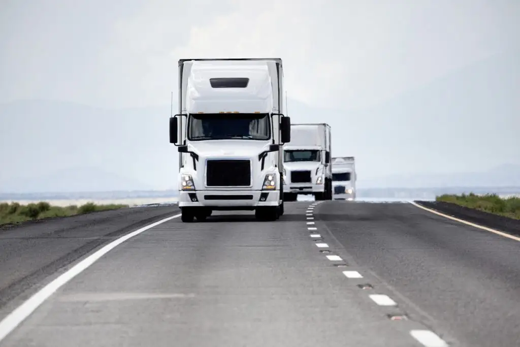 Three white semi-trucks driving on a highway in a mountainous landscape.