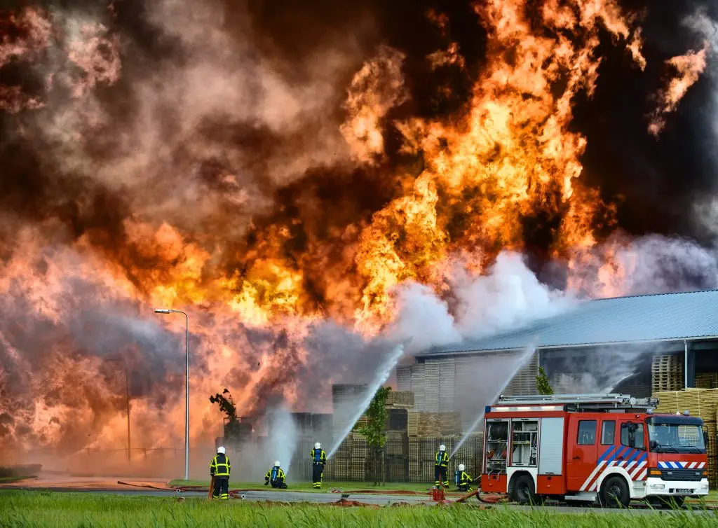 Firefighters using hoses to battle a large blaze at an industrial building with a fire truck in the foreground.
