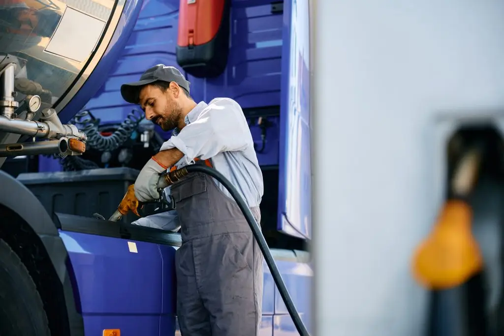 A man wearing a cap and overalls is refueling a large blue vehicle at a gas station.