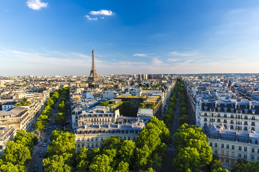 Aerial view of Paris featuring the Eiffel Tower, numerous buildings, and tree-lined streets under a clear blue sky.