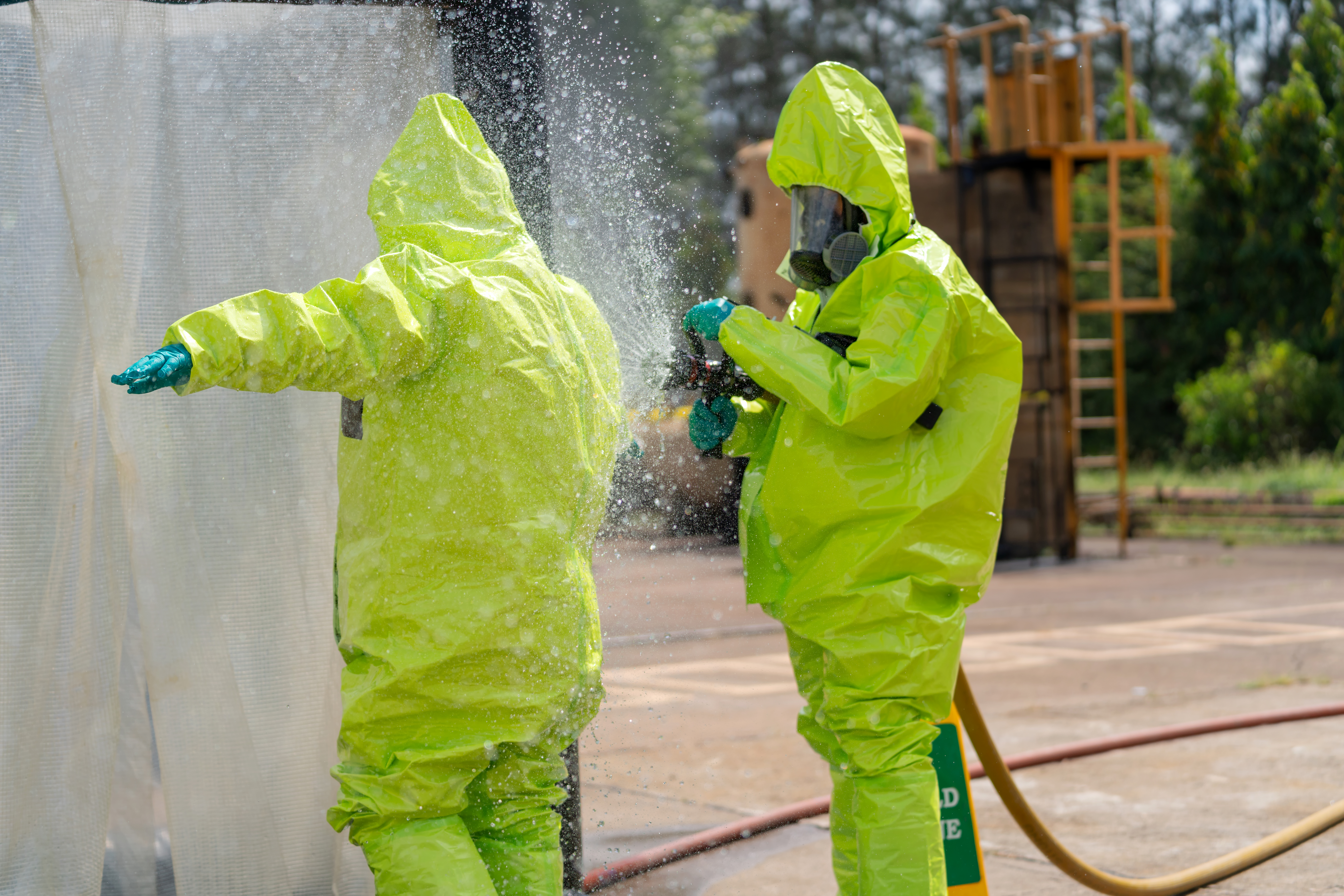 Two individuals in protective hazmat suits cleaning a surface with a high-pressure water spray outdoors.