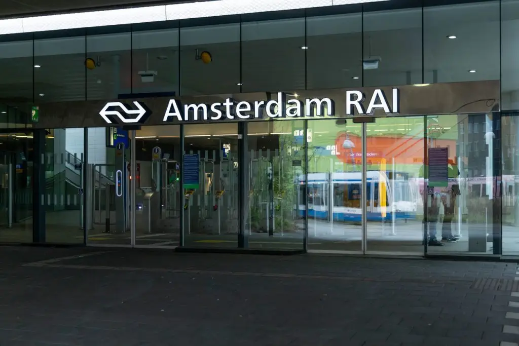 The entrance of the Amsterdam RAI train station is shown with glass doors and the station name. A moving tram is visible in the reflection behind the glass.
