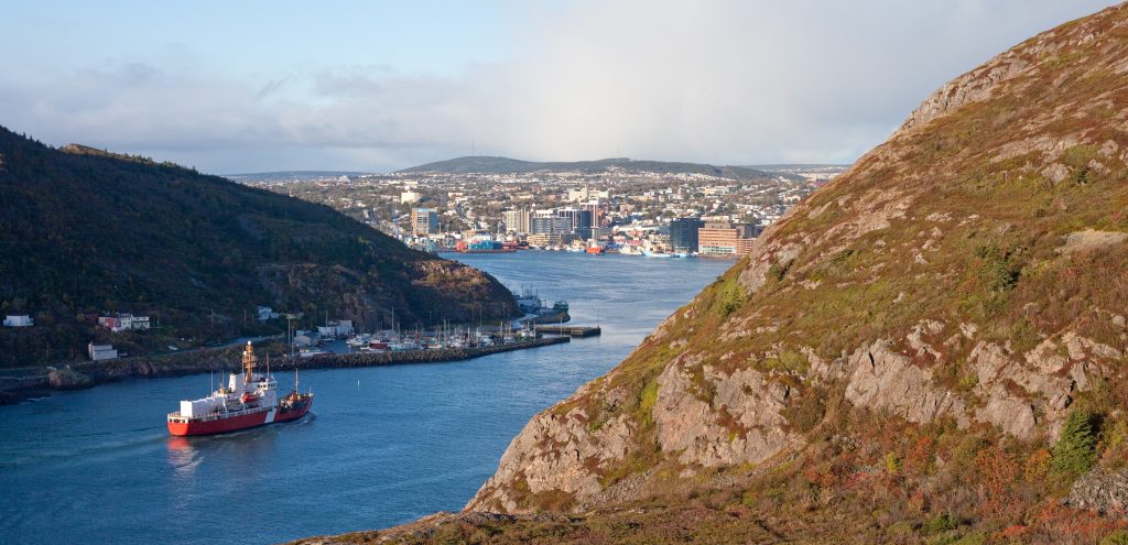 A red ship sails through a narrow harbor passage flanked by hills, with a city and its waterfront visible in the background under a partly cloudy sky.