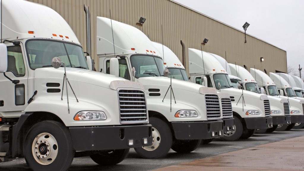 A row of white semi-trucks is lined up in front of a large warehouse on an overcast day.