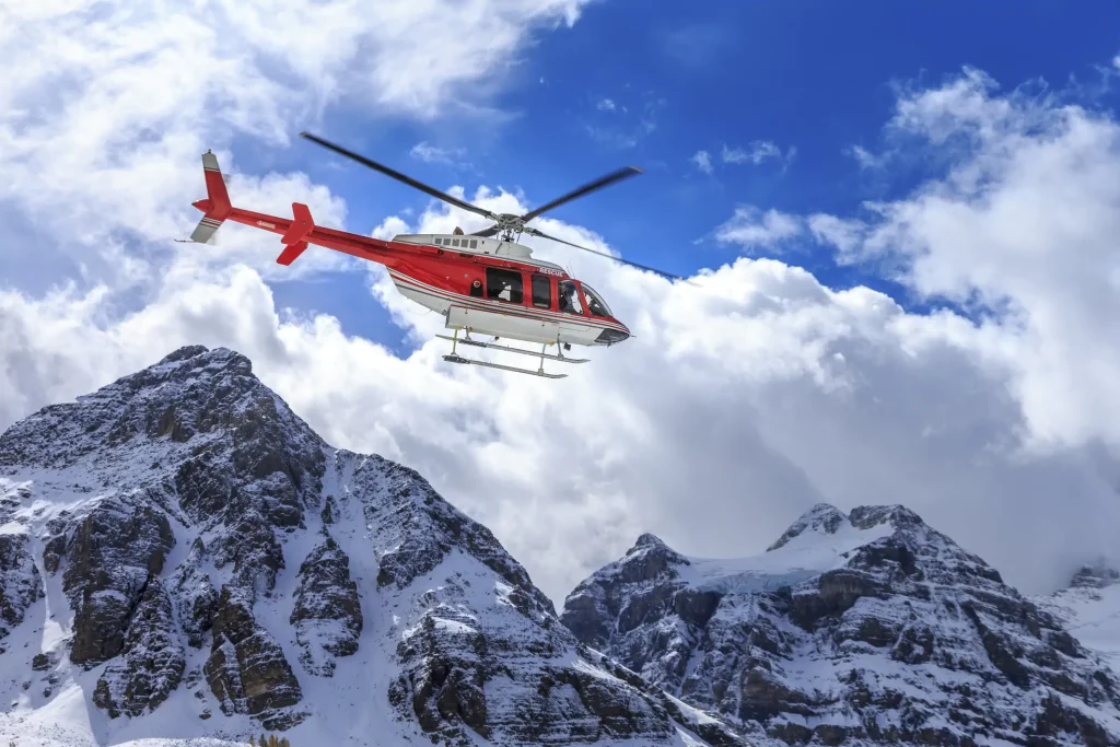 A red helicopter flying in a clear blue sky near snow-covered mountain peaks.